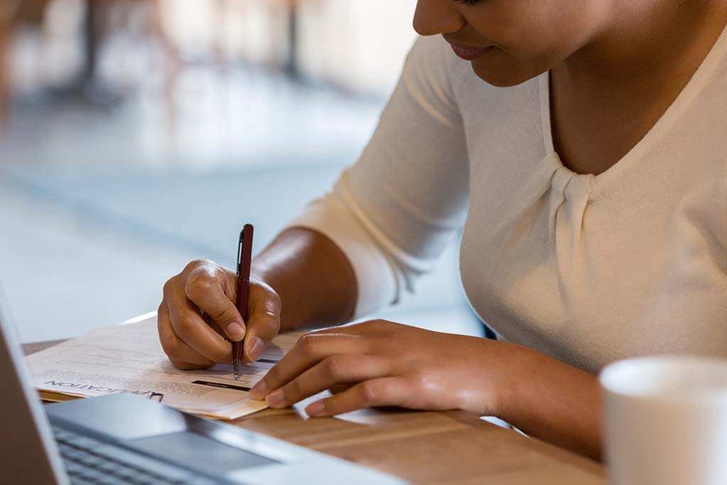 Woman filling out a form with pen and paper in front of a laptop